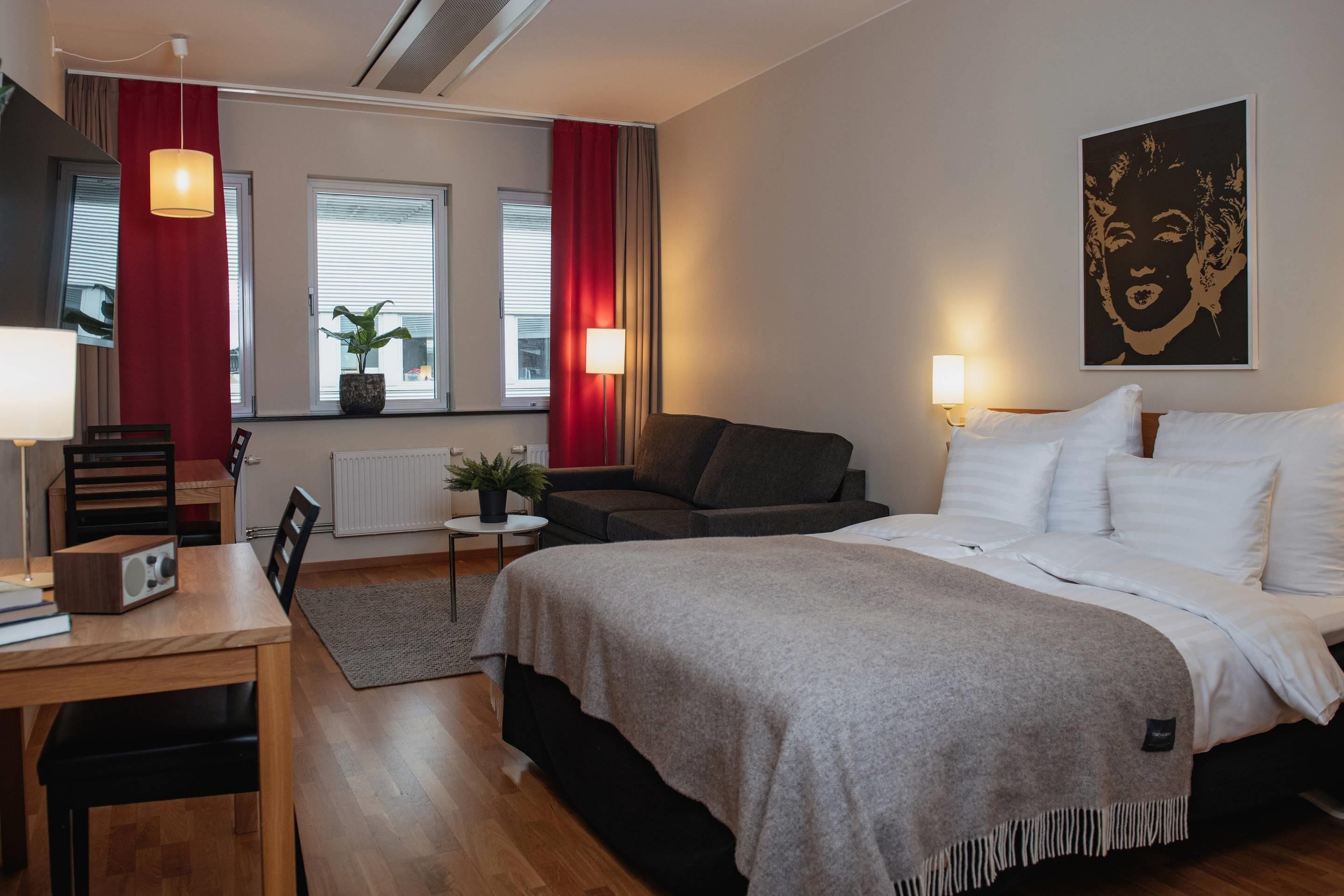 Habitación de hotel con cama, escritorio, silla, una mesita, planta en maceta y ventana con cortinas.