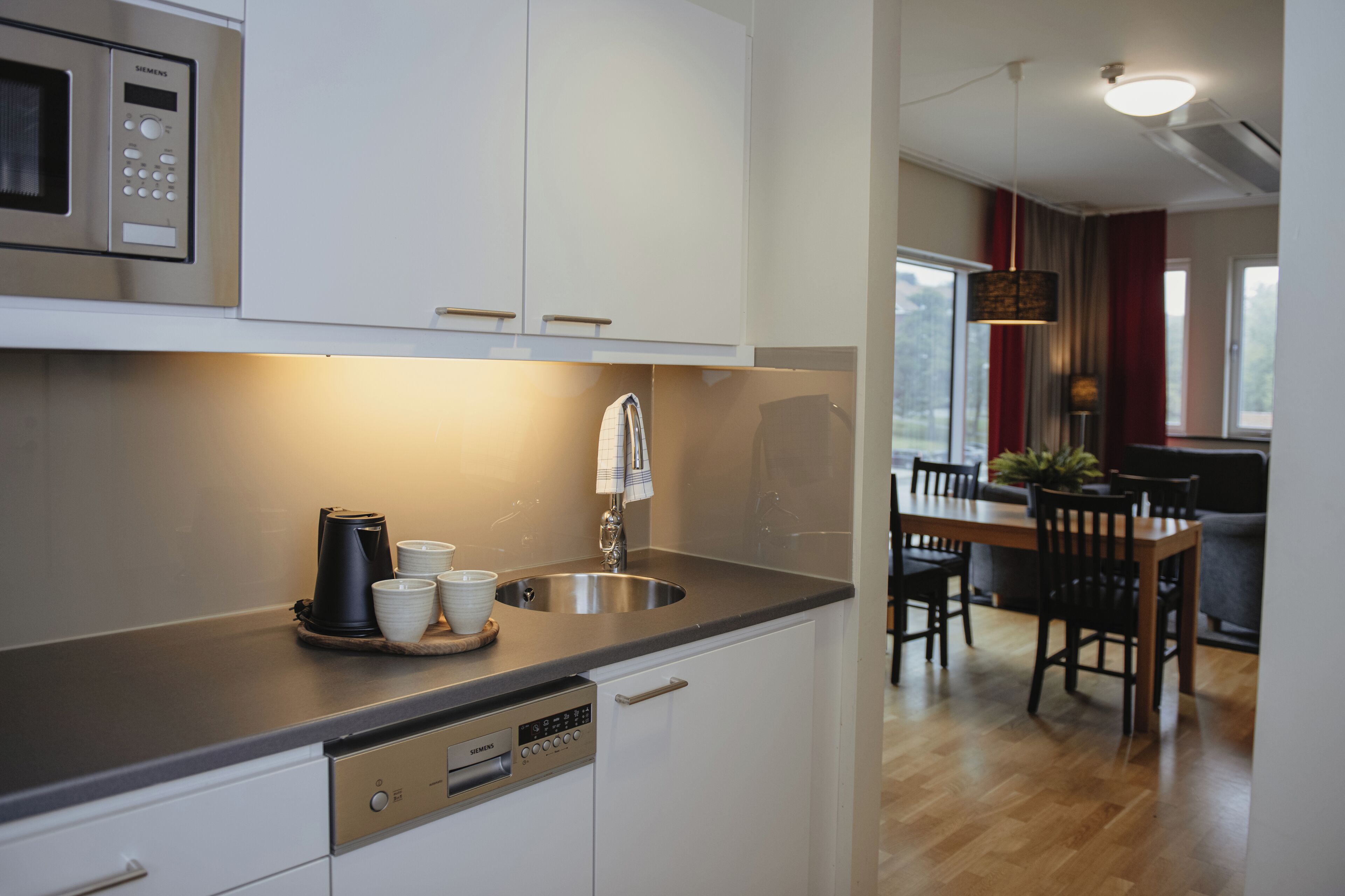 Una cocina moderna con armarios blancos, un lavamanos de acero inoxidable y una cafetera. Se observa un comedor con una mesa y sillas de madera al fondo.