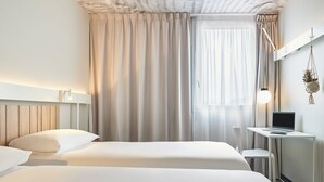 Ein Hotelzimmer mit zwei Betten, einem Schreibtisch mit einem Laptop, einer hängenden Pflanze und einem großen Fenster mit Vorhängen.