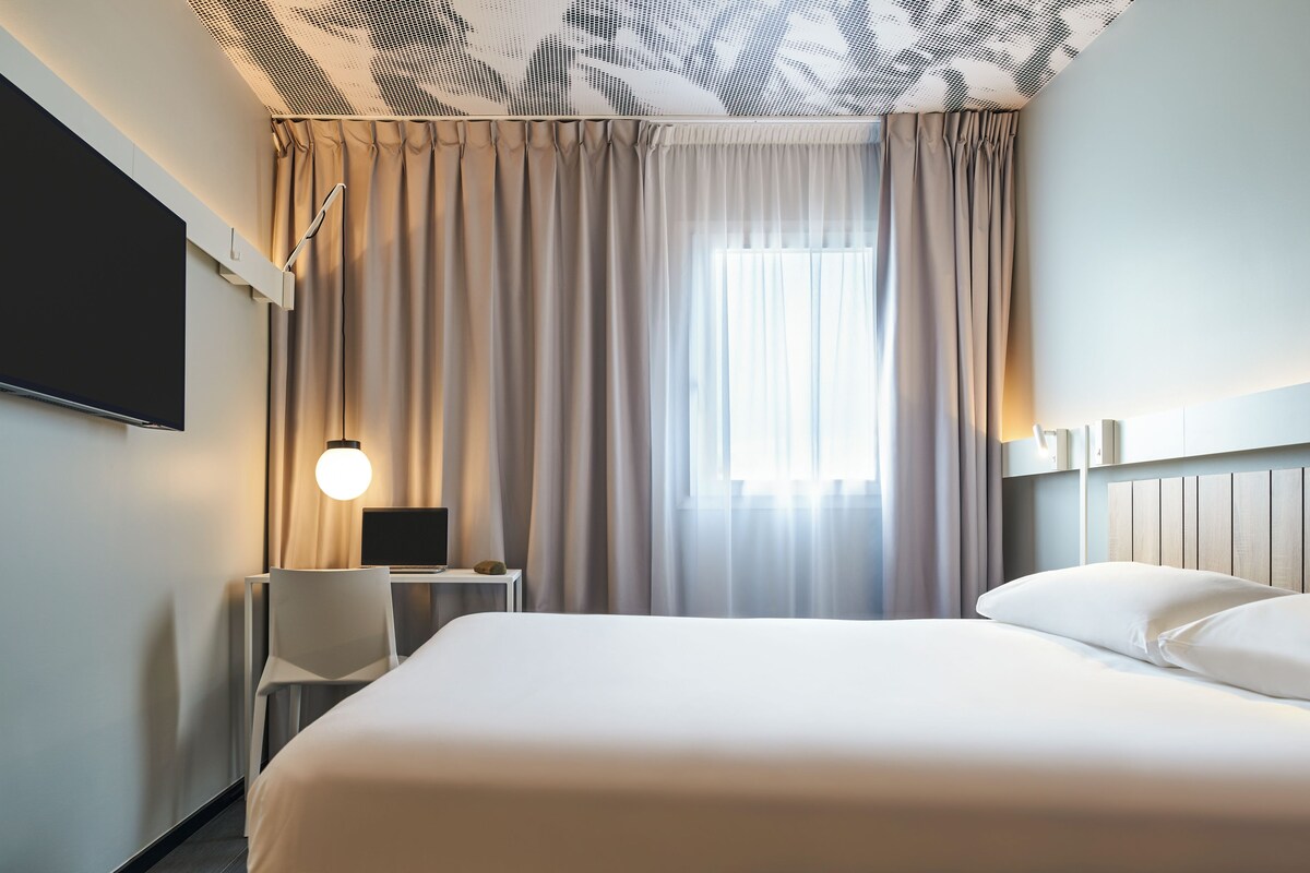 Ein Hotelzimmer mit Bett, Schreibtisch, Stuhl, Fernseher und einem Fenster mit Vorhängen.