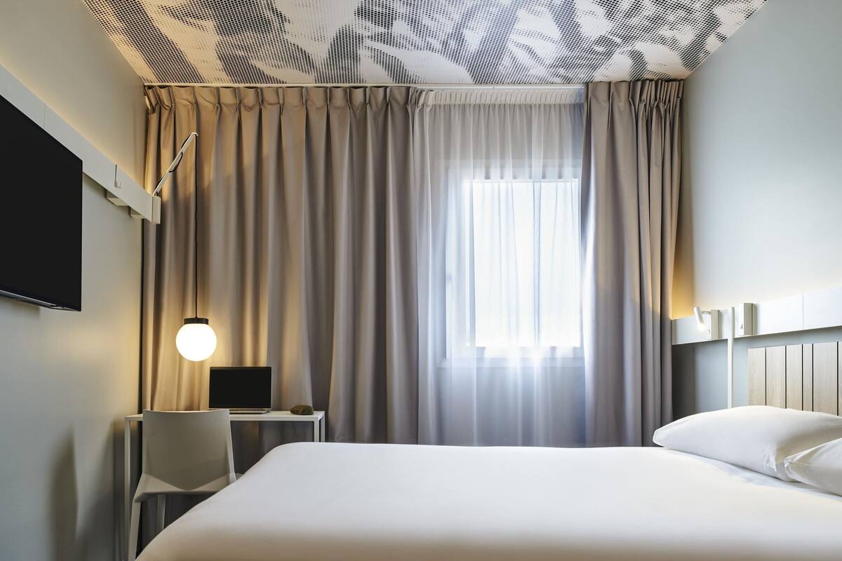 A hotel room with a bed, a desk, a chair, a TV, and a window with curtains.