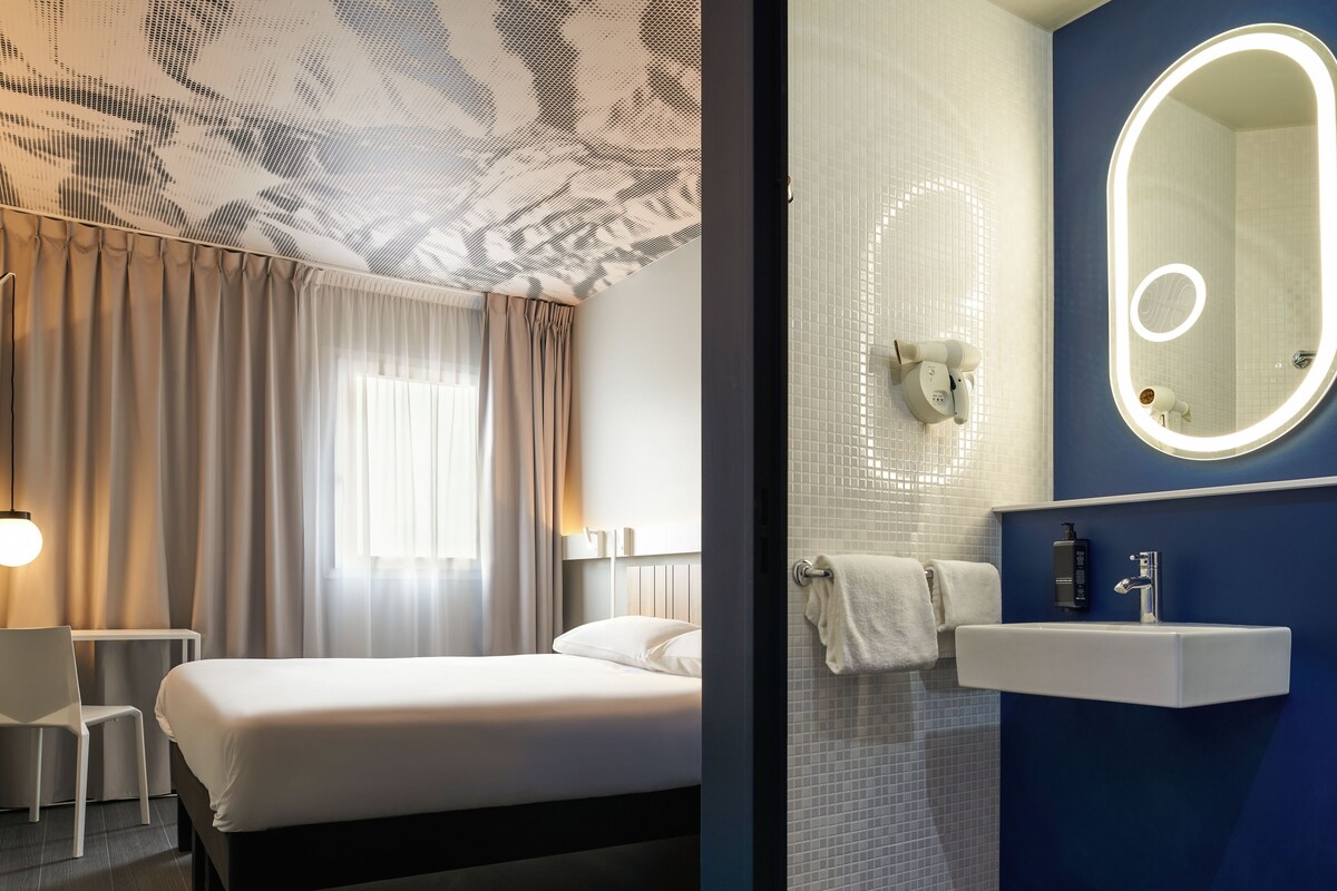 Ein Hotelzimmer mit Bett, Schreibtisch, Stuhl und einem Badezimmer mit Waschbecken und Spiegel.