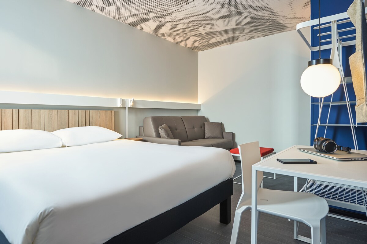 Ein modernes Hotelzimmer mit einem großen Bett, einem Schreibtisch mit Stuhl, einer Couch und einer Deckenleuchte.