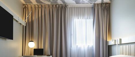 Habitación de hotel con cama, escritorio, silla, televisor y ventana con cortinas.