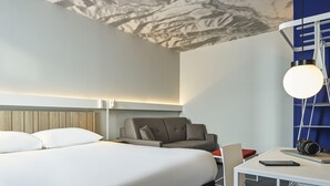 Une chambre d’hôtel moderne avec un grand lit, un bureau avec un ordinateur portable, une chaise et une petite table sur laquelle se trouve un appareil photo.
