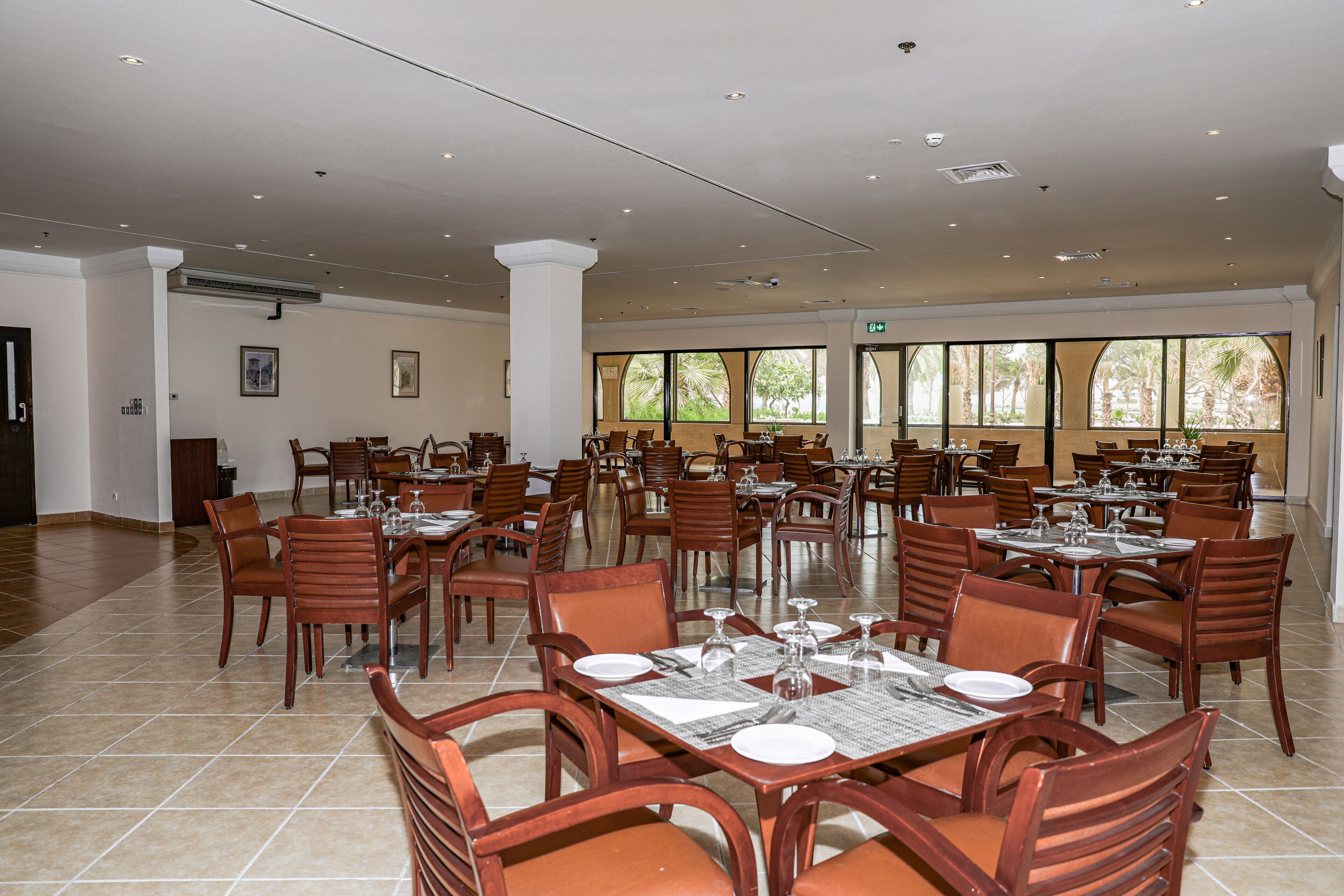 A spacious dining area with multiple tables and chairs, large windows with views of palm trees, and a tiled floor.