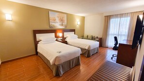 A hotel room with two beds, a painting on the wall, a desk with a chair, and a large window with curtains. - Dhafra Beach Hotel (Jebel Dhanna)