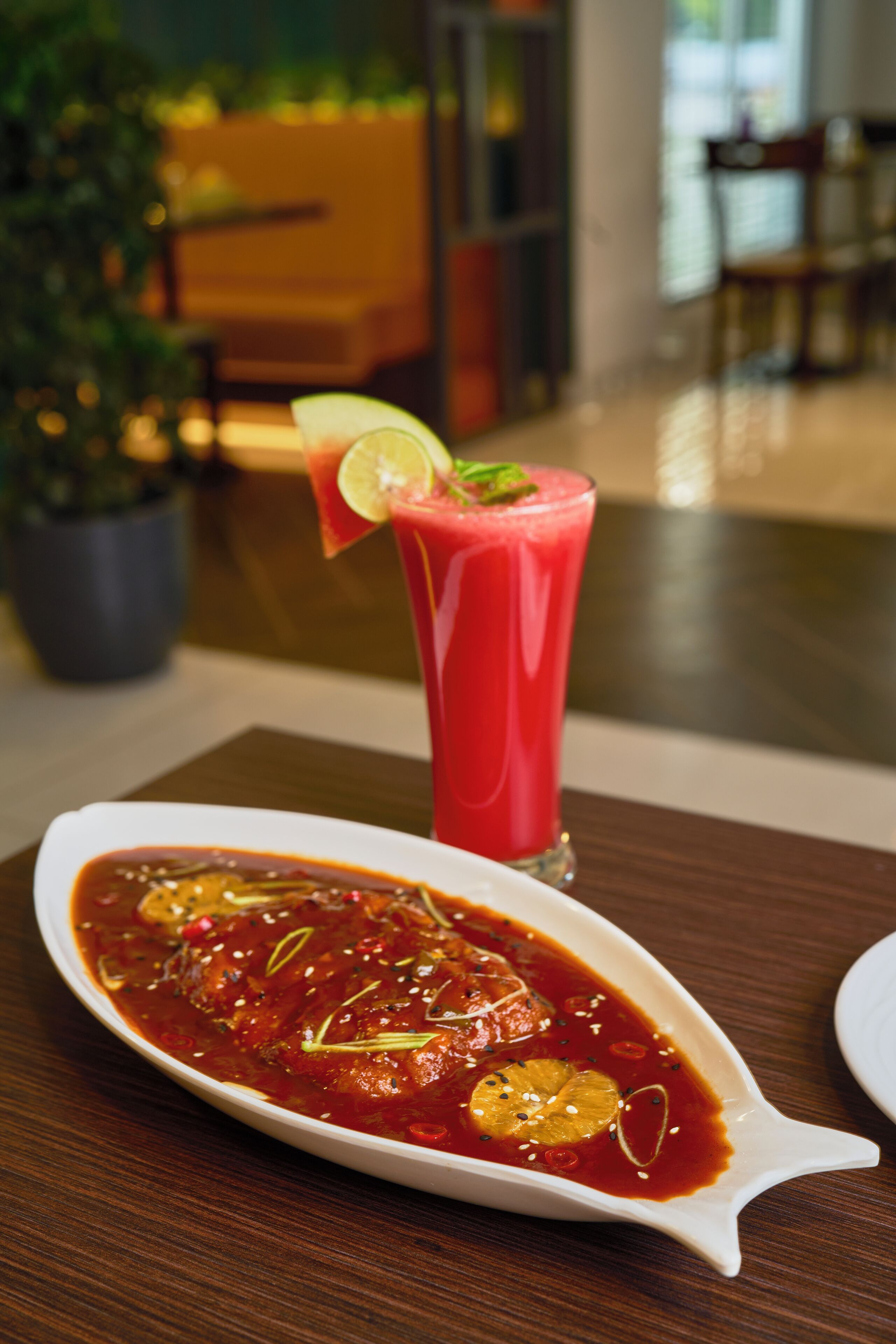 Una mesa de comedor con un plato blanco ovalado que contiene un guiso de salsa roja adornado con hierbas y rodajas de limón, y un vaso alto de bebida roja con una rodaja de limón como adorno.
