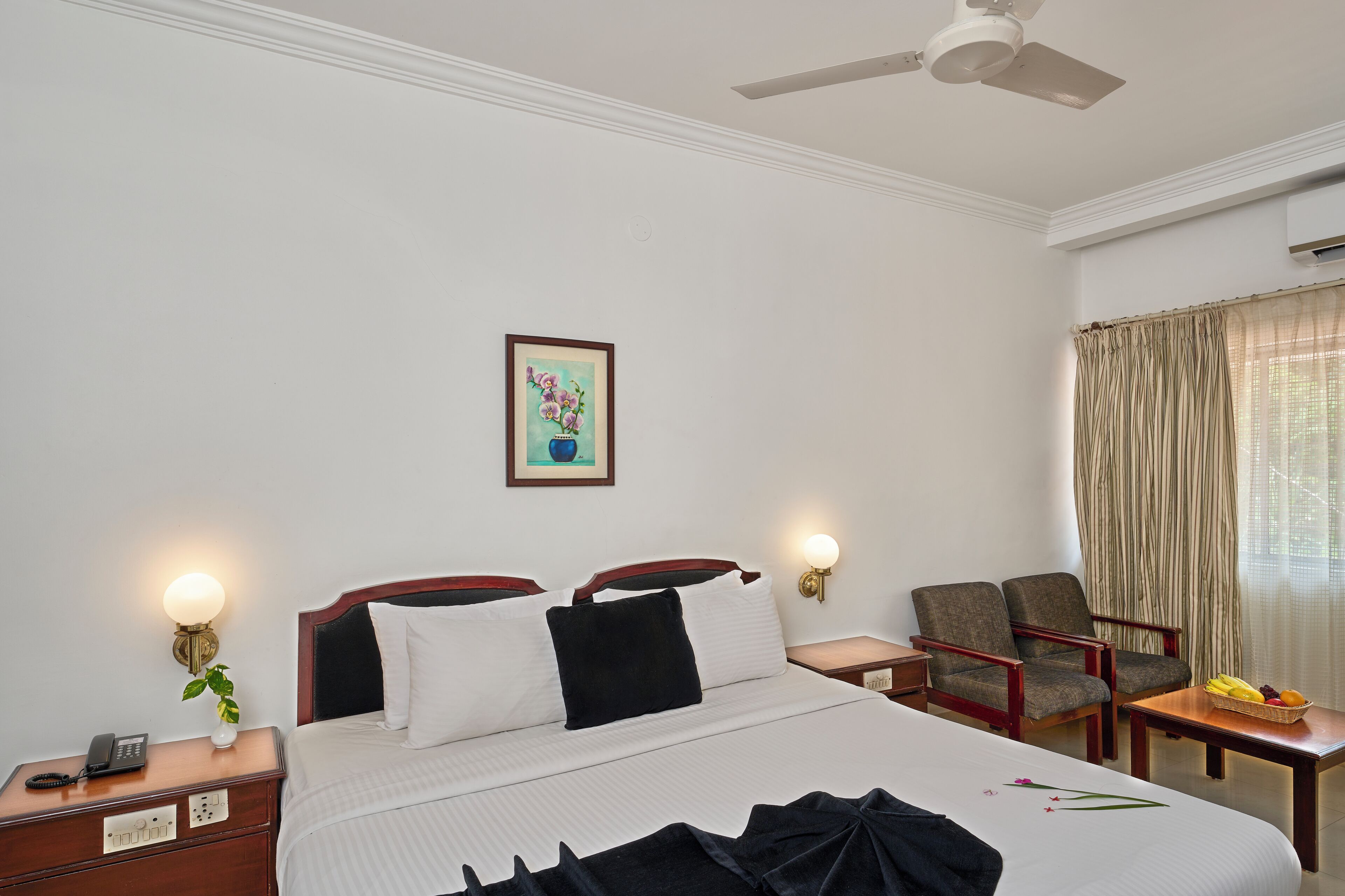 A hotel room with a bed, bedside tables, a chair, a small table with fruit, and a painting on the wall.