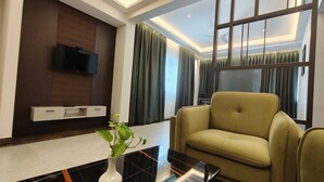 Una sala de estar moderna con un sofá, un televisor de pantalla plana montado en un panel de madera, un jarrón con una planta y ventanas grandes con cortinas.