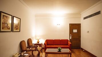 Een woonkamer met een houten vloer, een rode bank, een salontafel en een lamp. Er hangen afbeeldingen in lijsten aan de muur en op een bijzettafeltje staat een telefoon.