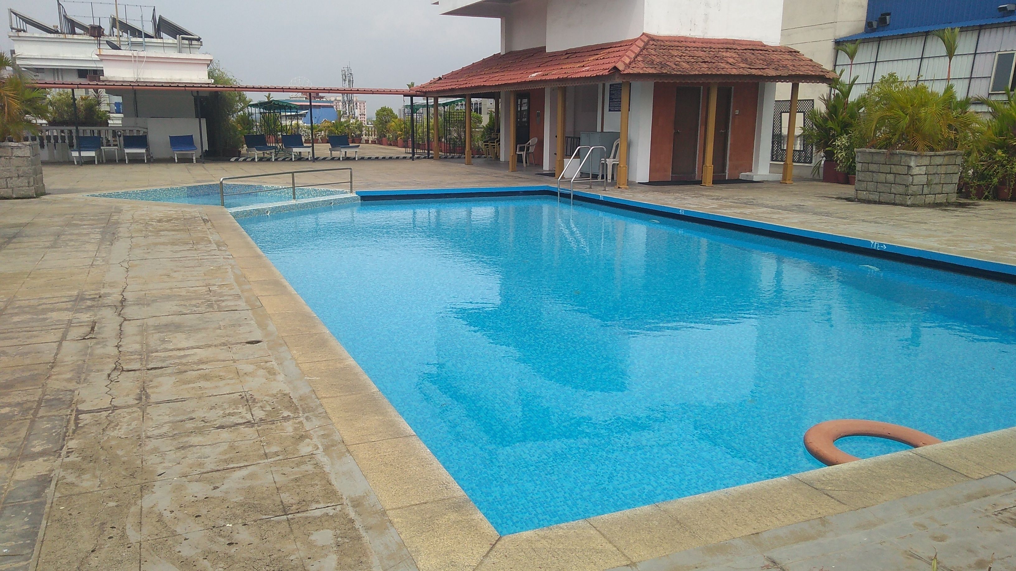 Une piscine rectangulaire entourée d’une terrasse carrelée, un petit espace de repos couvert et, au fond, un bâtiment avec un toit rouge.