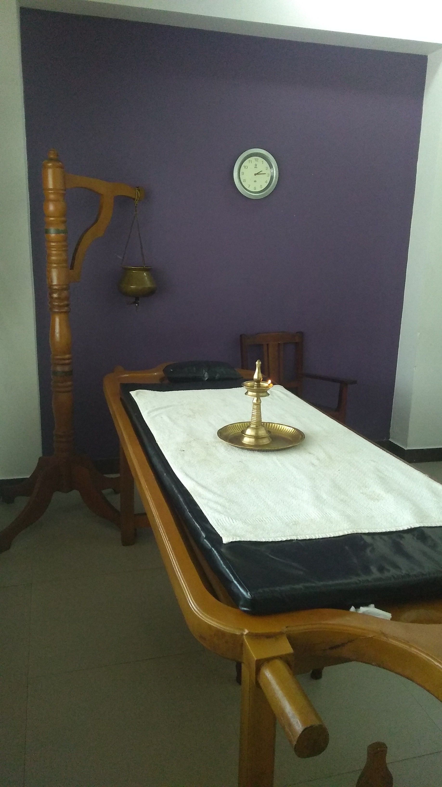 A room with a wooden table, a brass lamp, a clock on the wall, and a wooden chair.