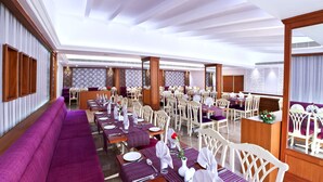 Uma ampla sala de jantar com várias mesas preparadas para o serviço, um extenso sofá roxo e um teto alto com iluminação natural.