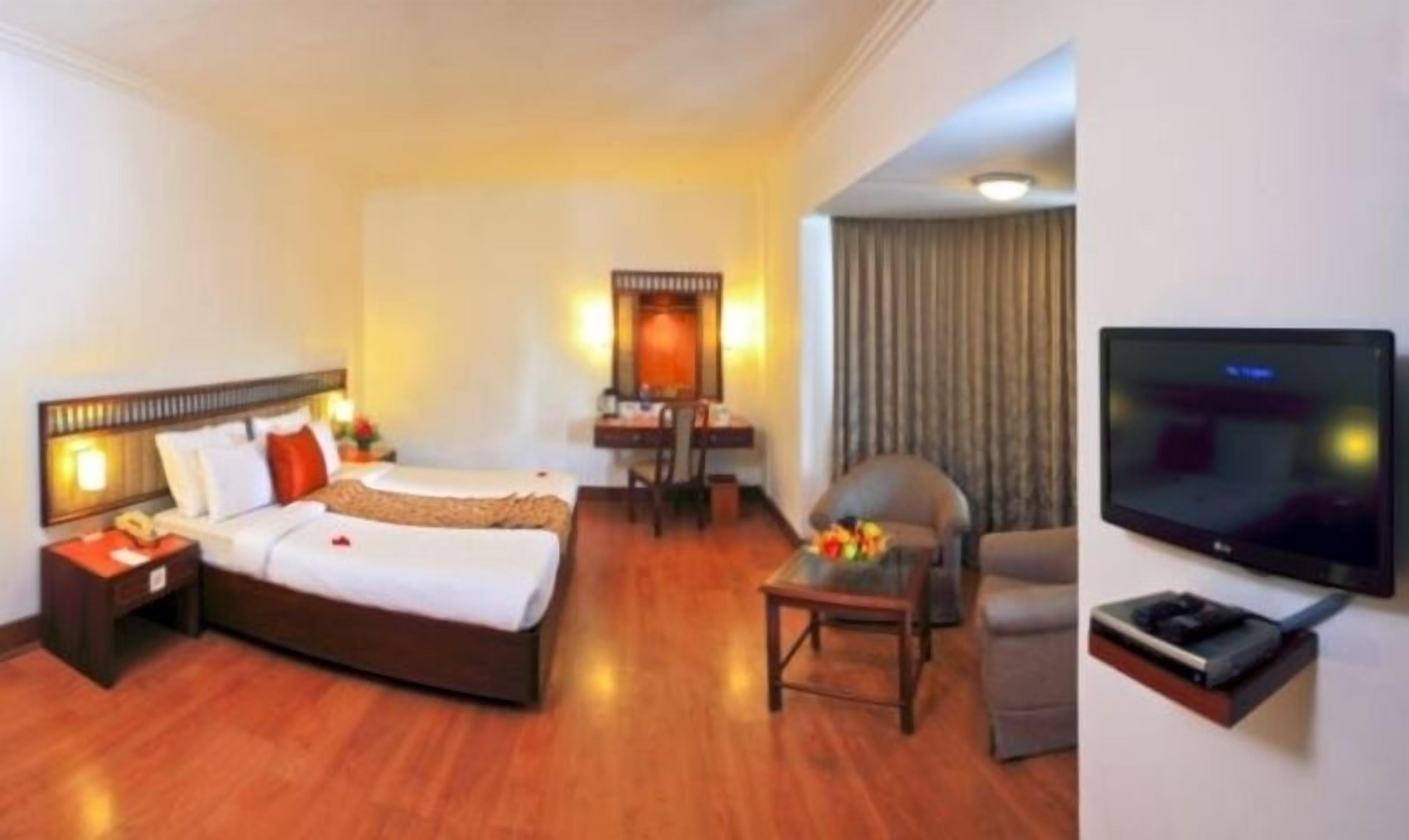 Quarto de hotel com cama, escrivaninha, cadeira, televisão e uma mesinha com flores.