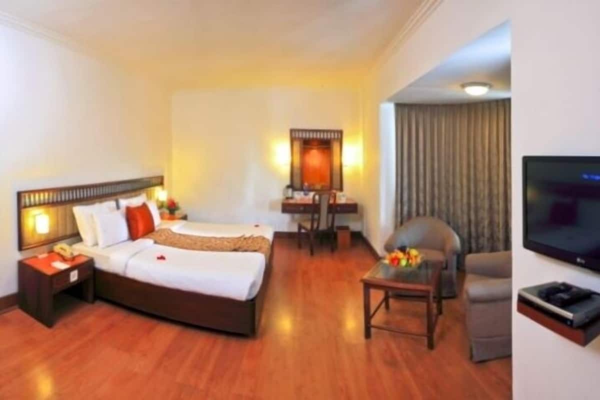Quarto de hotel com cama, escrivaninha, cadeira, televisão e uma mesinha com flores.