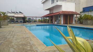 A swimming pool with a tiled deck, a covered seating area, and a building with a red roof in the background.