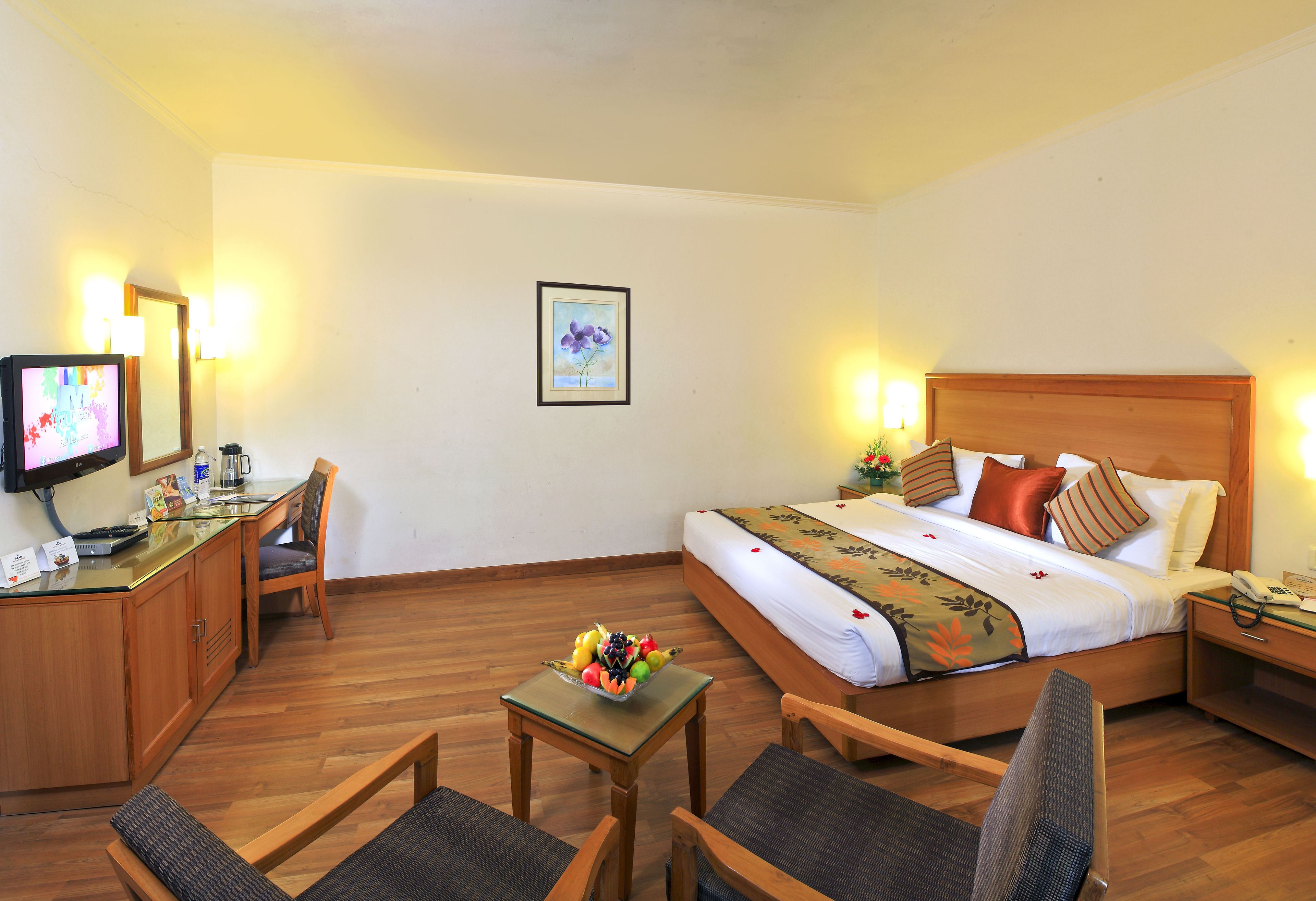 A hotel room with a bed, a desk with a computer, a chair, a small table with fruit, and a bedside table with a lamp.