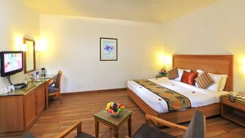 A hotel room with a bed, a desk with a computer, a chair, a small table with fruit, and a bedside table with a lamp.