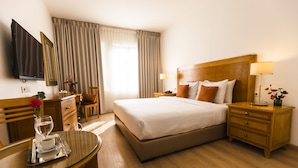 A hotel room with a large bed, a bedside table, a TV mounted on the wall, a desk with a chair, and a window with curtains. - Liwan Hotel (Amman)