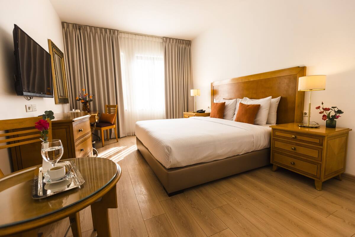 A hotel room with a large bed, a bedside table, a TV mounted on the wall, a desk with a chair, and a window with curtains.