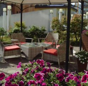 Un'area salotto esterna con sedie in vimini e un tavolo in vetro, circondata da vasi con piante e fiori.