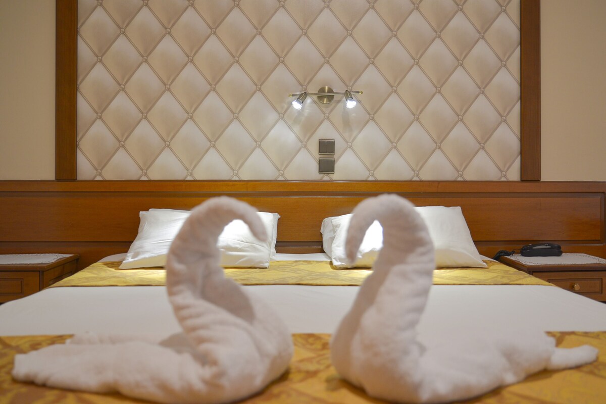 A hotel room with a bed, two swan-shaped pillows, a nightstand, a lamp, and a wall with a diamond-patterned design.