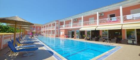 A swimming pool with lounge chairs, a covered area with umbrellas, and a building with balconies and a storefront.