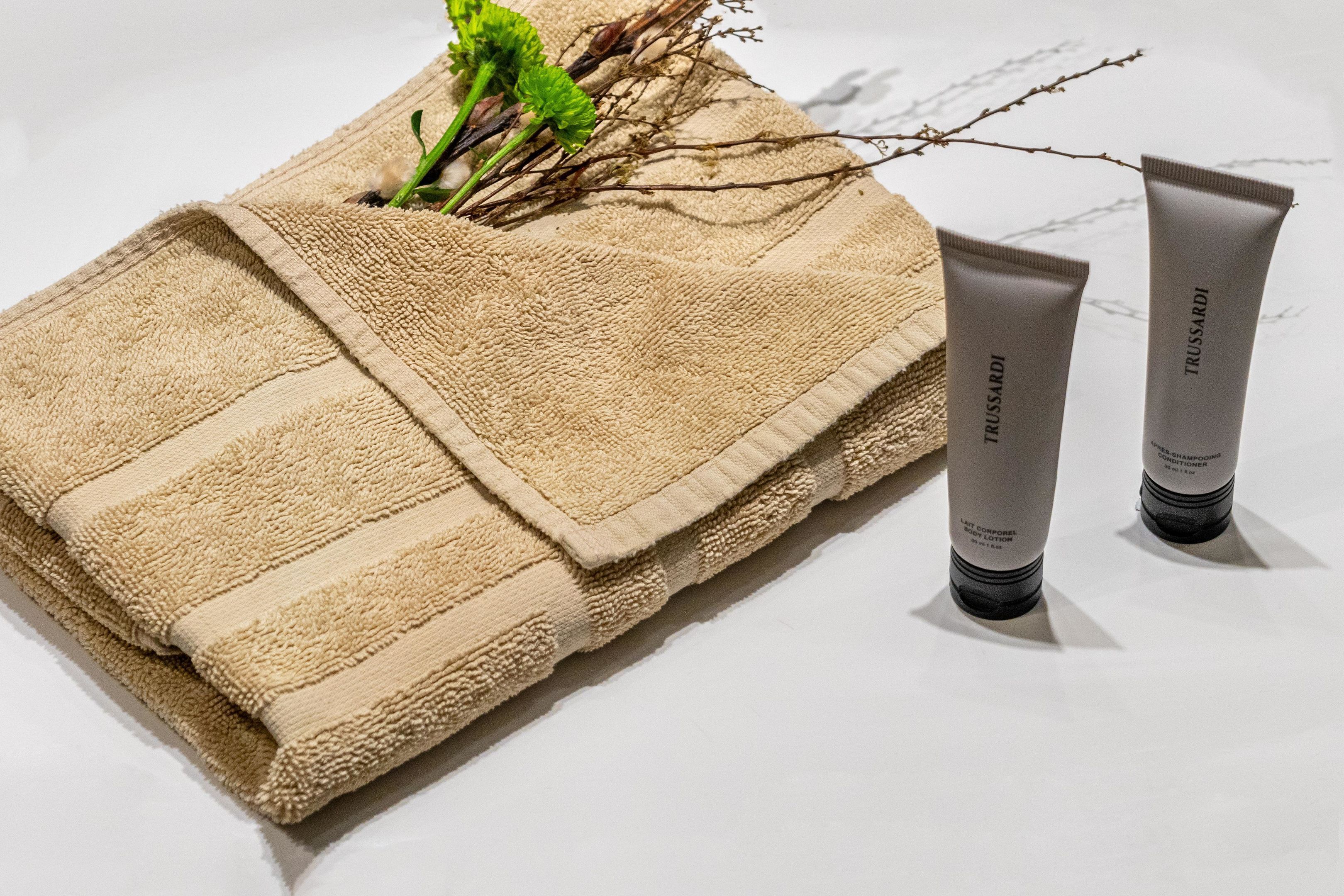 Ein beiges Handtuch mit Strukturmuster, zwei Tuben Trussardi Körperlotion und ein Zweig mit grünen Blättern.