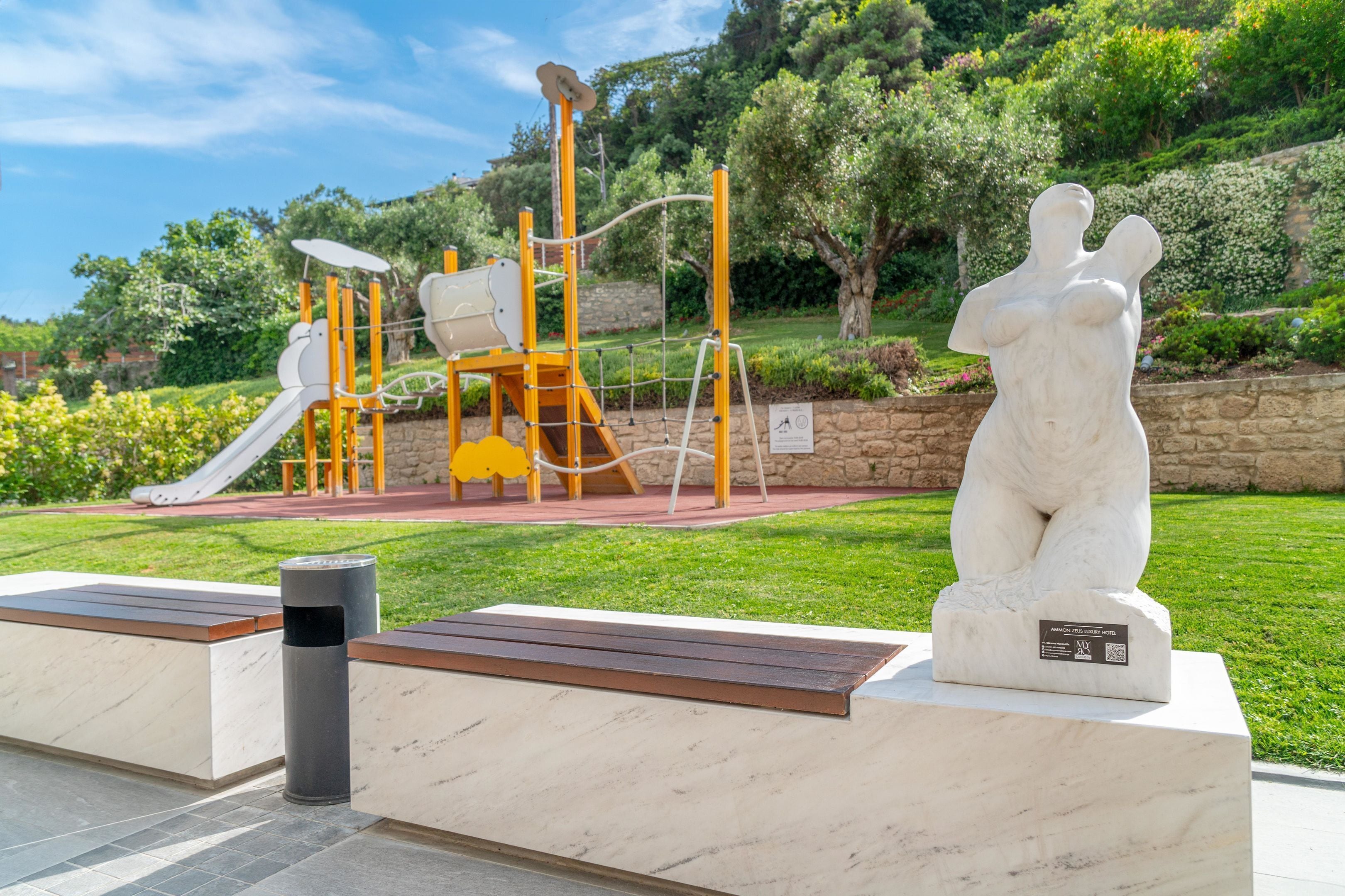 A playground with a slide and climbing structures, surrounded by greenery and a stone wall.