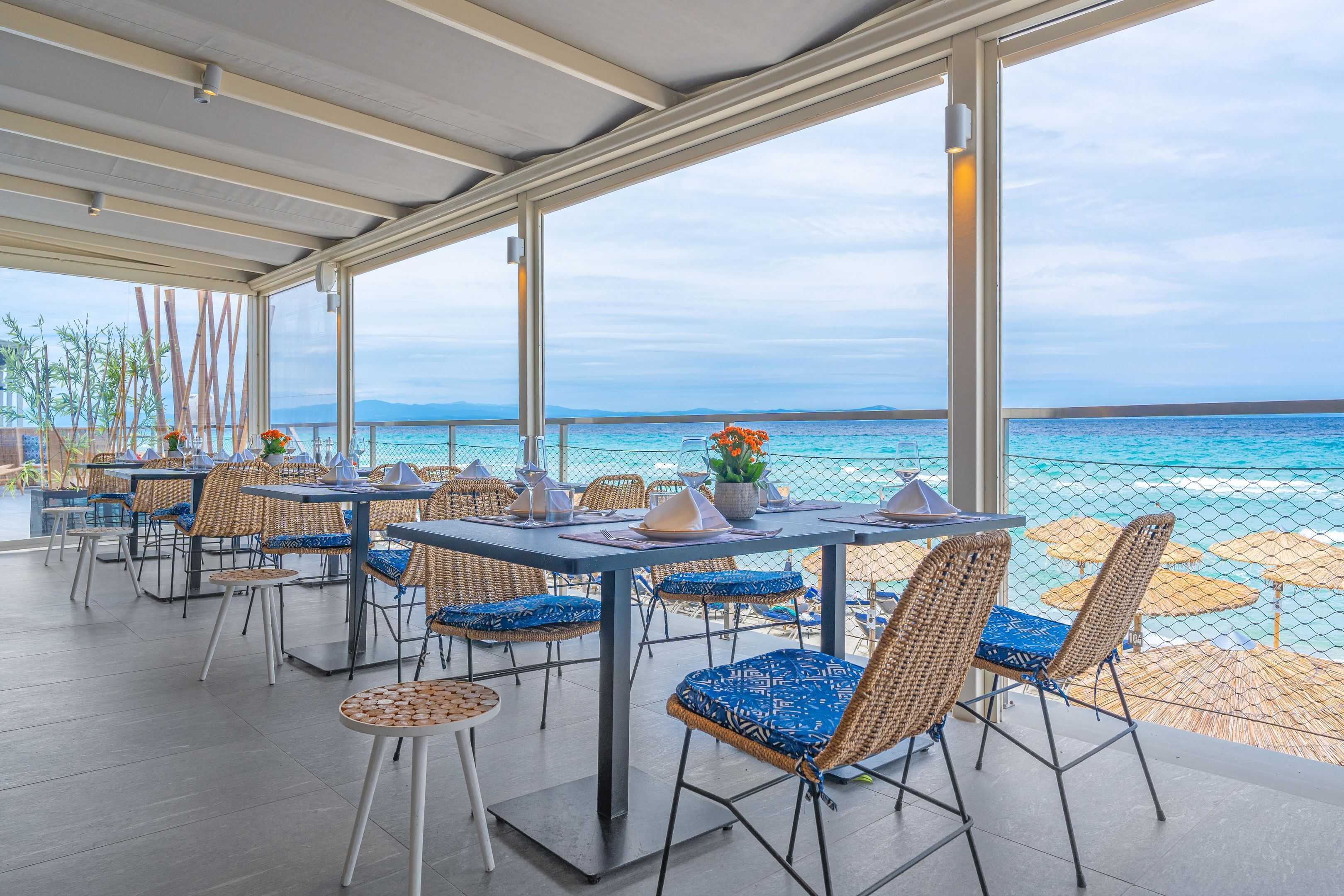 An outdoor dining area with tables and chairs, overlooking a beach and the sea.