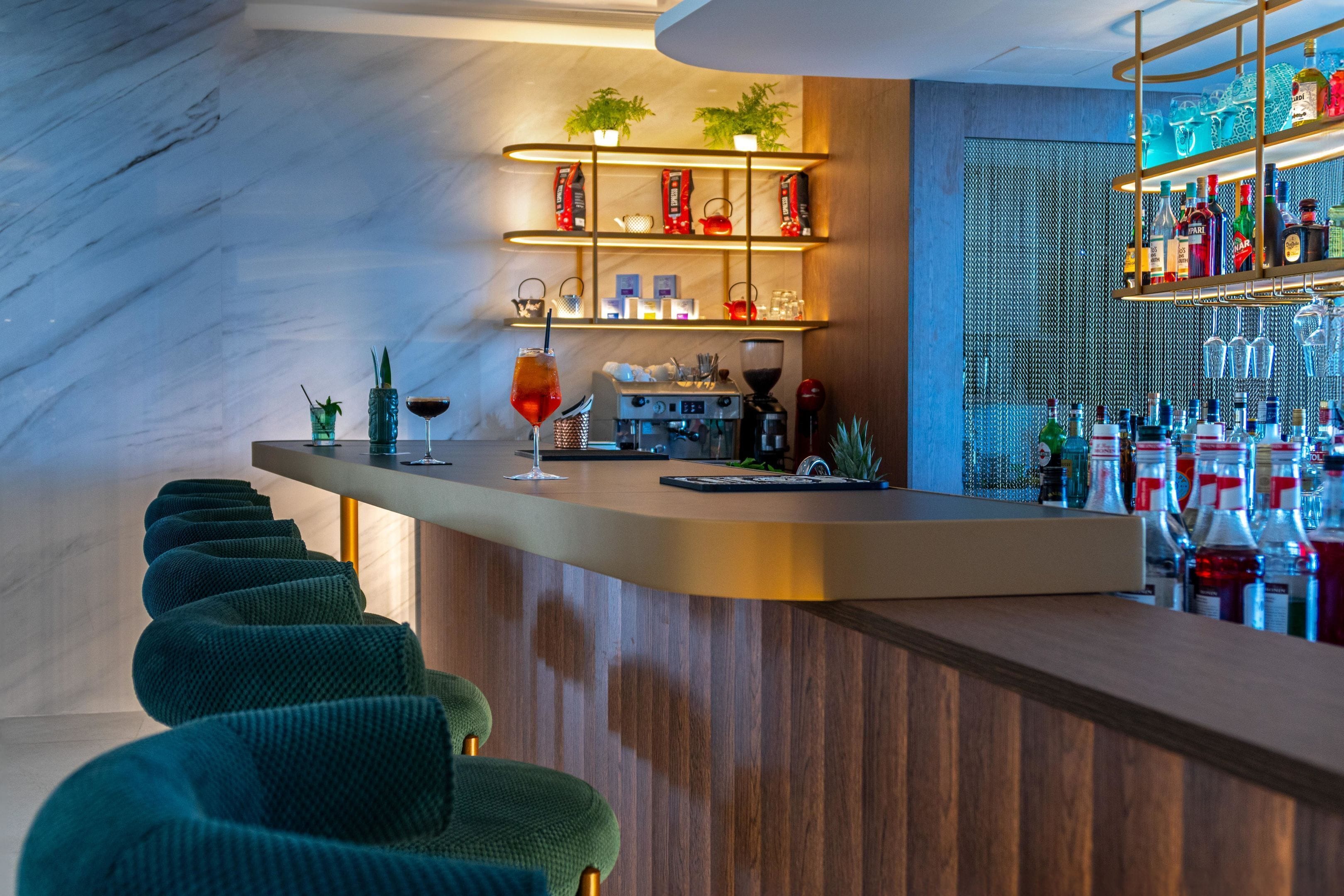 Un espace bar doté d’un comptoir en bois, de tabourets de bar recouverts d’un tissu vert, d’étagères remplies de bouteilles et de verres, et d’un mur en marbre.