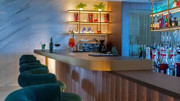 Un espace bar doté d’un comptoir en bois, de tabourets de bar recouverts d’un tissu vert, d’étagères remplies de bouteilles et de verres, et d’un mur en marbre.