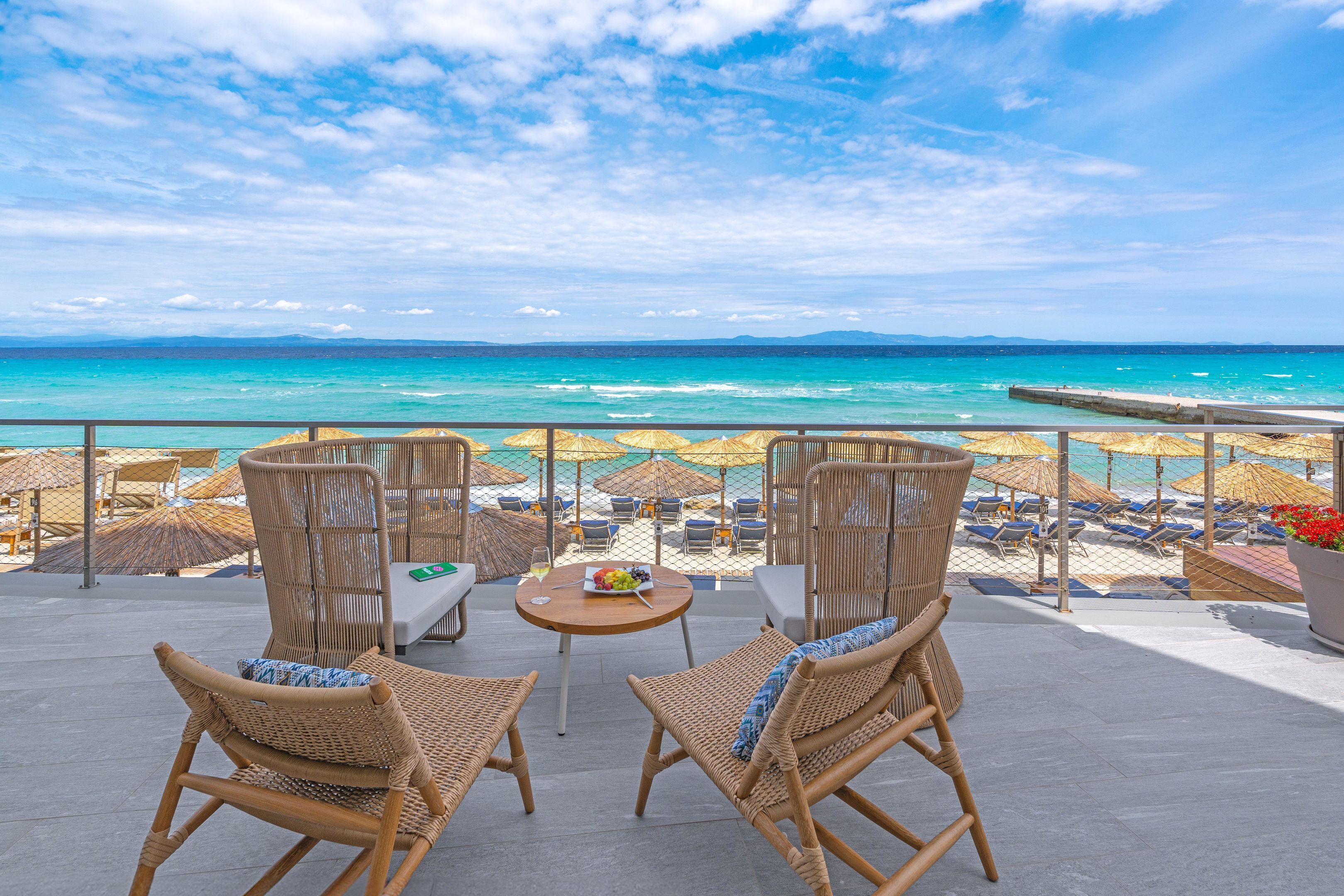 A terrace with wicker chairs and a table, overlooking a beach with umbrellas and lounge chairs.