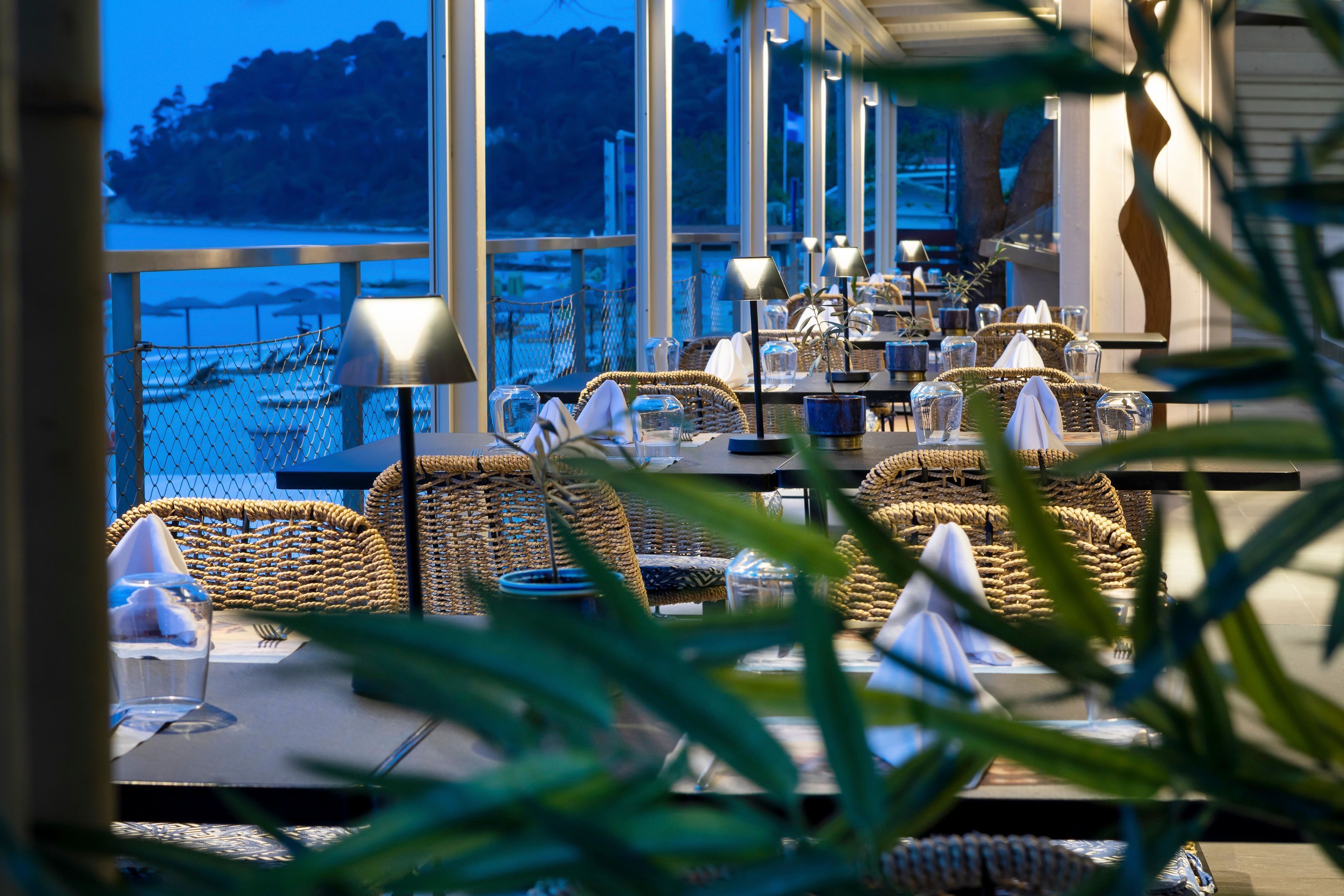 A restaurant with wicker furniture, tables set with glassware, and a view of the water.