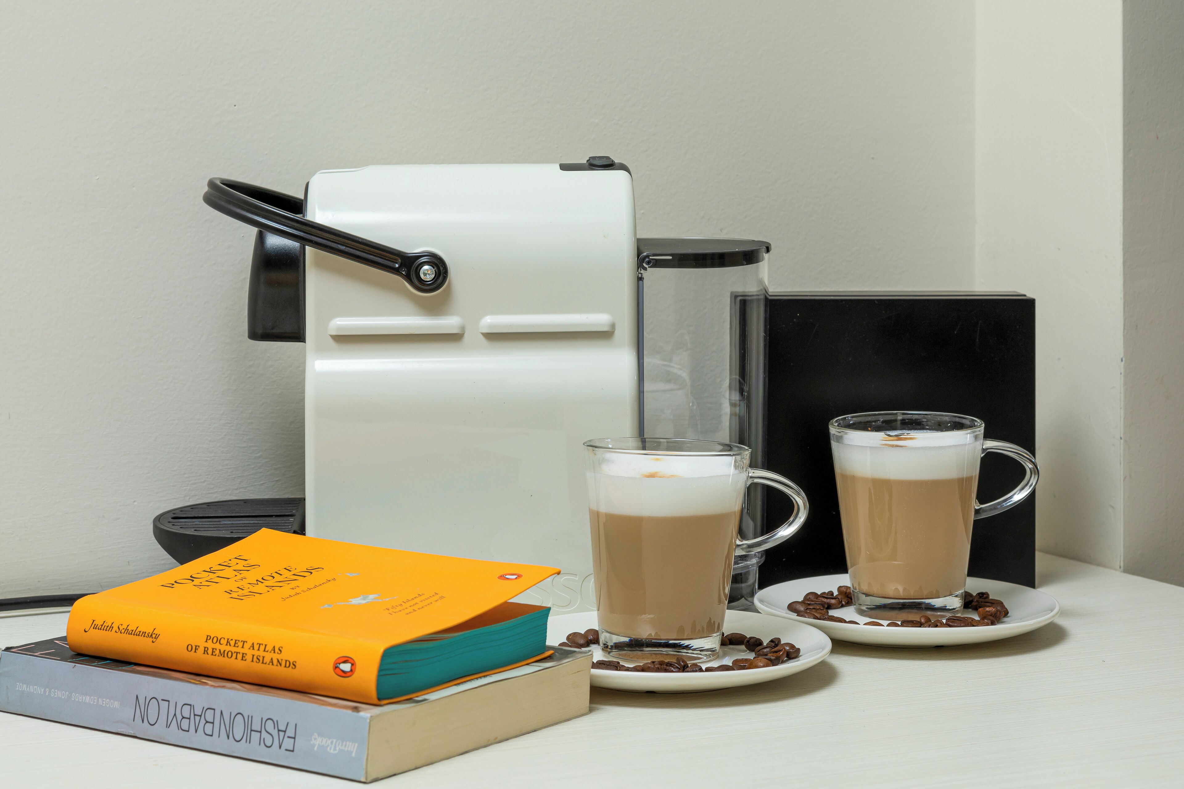 A coffee maker, two cups of coffee, a book titled 'Pocket Atlas of Remote Islands', and a coffee pod.