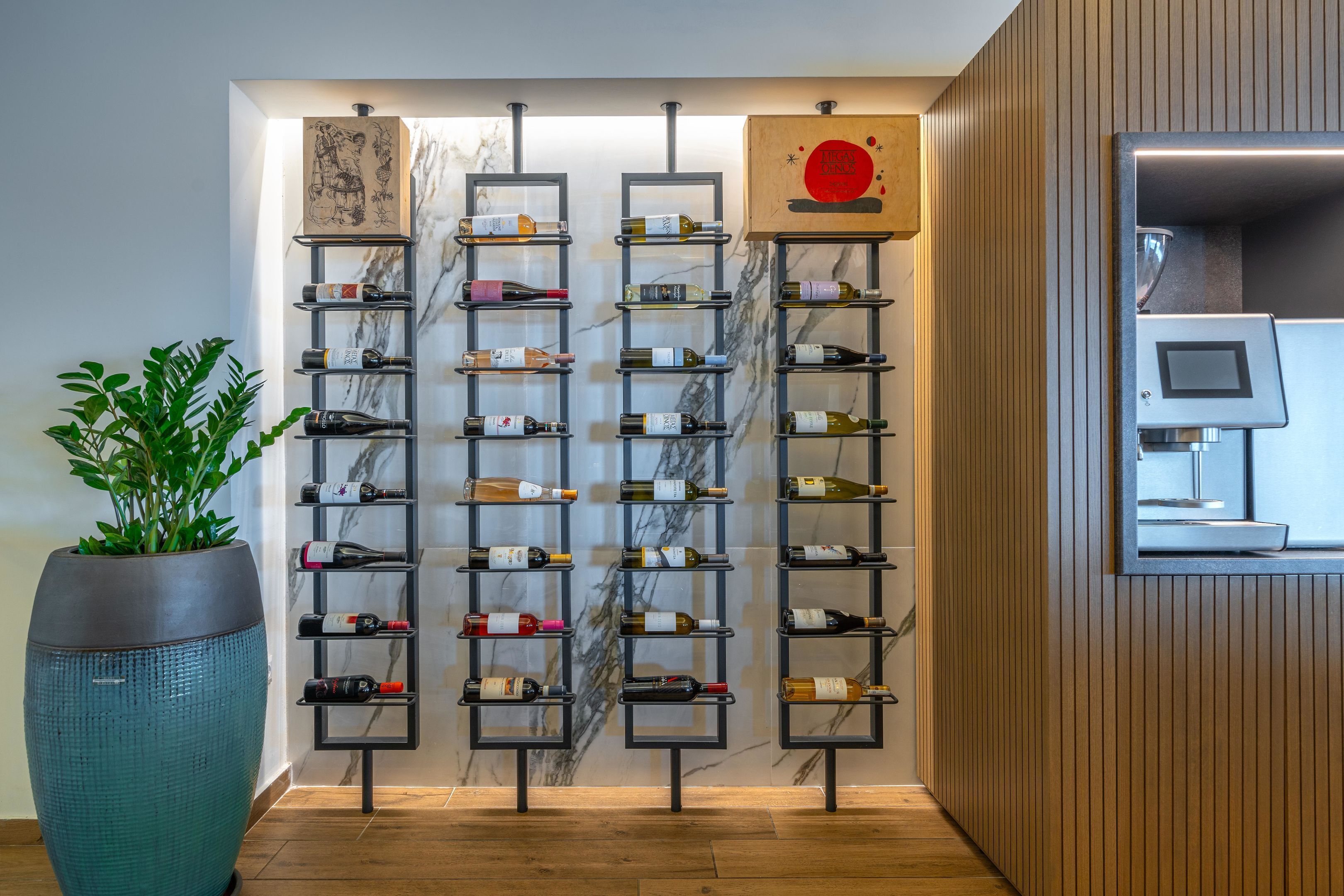 A modern hotel lobby with a wine rack displaying various bottles, a large potted plant, and a wooden wall panel.