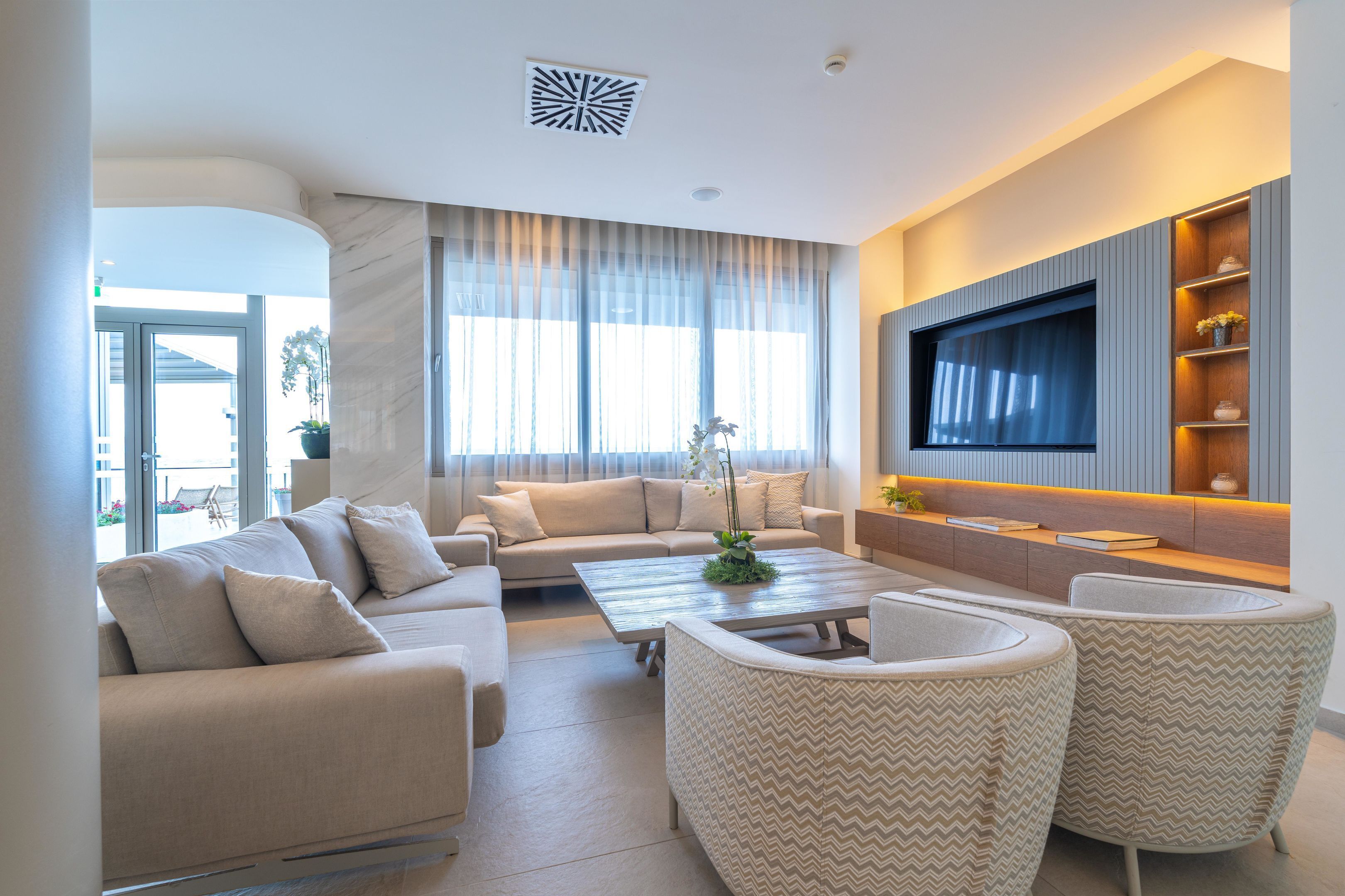 A modern living room with a sofa, coffee table, and armchairs, a flat-screen TV mounted on the wall, and a large window with sheer curtains.