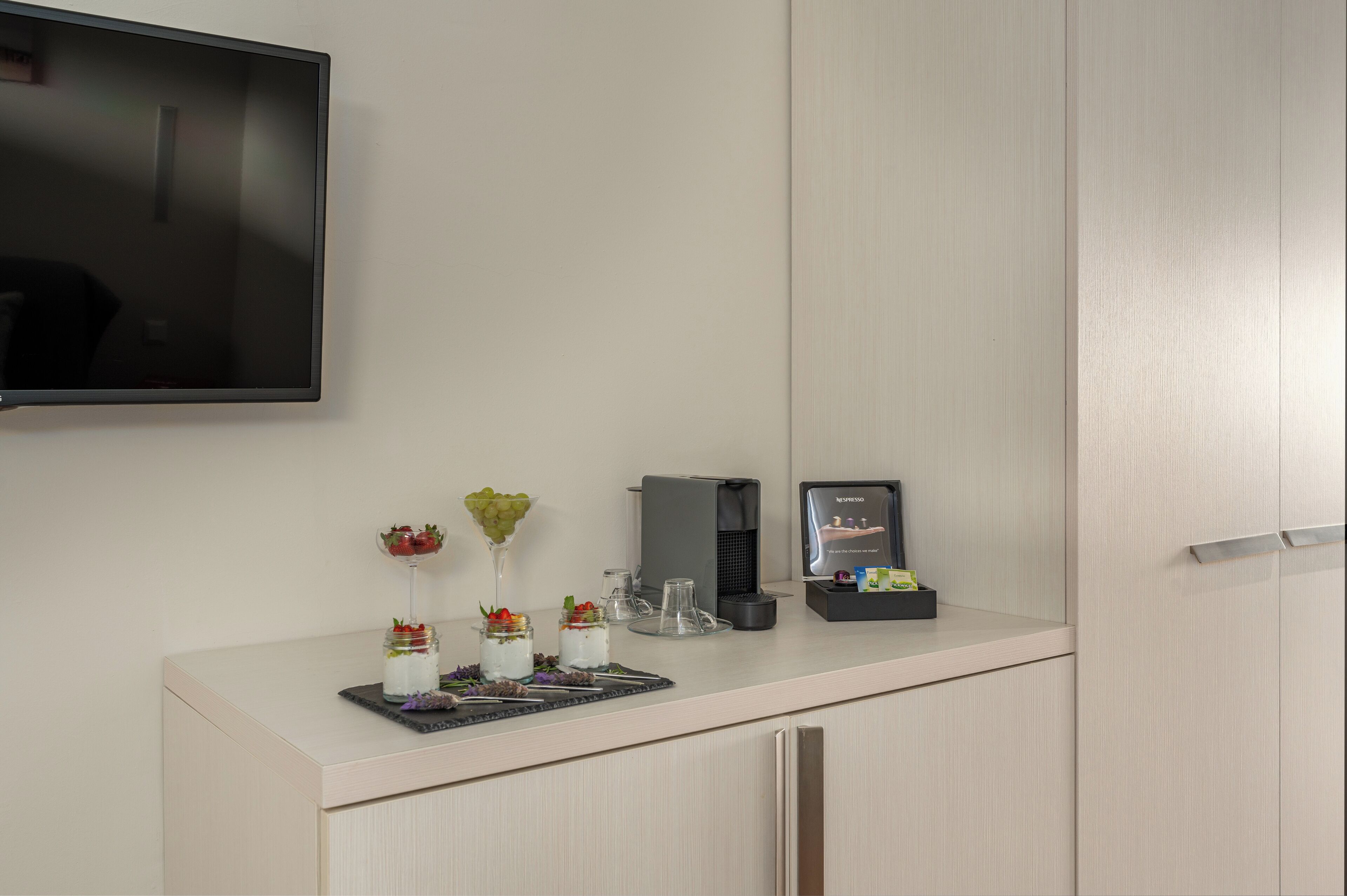 A modern kitchen with a flat-screen TV mounted on the wall, a coffee machine, and a small refrigerator.