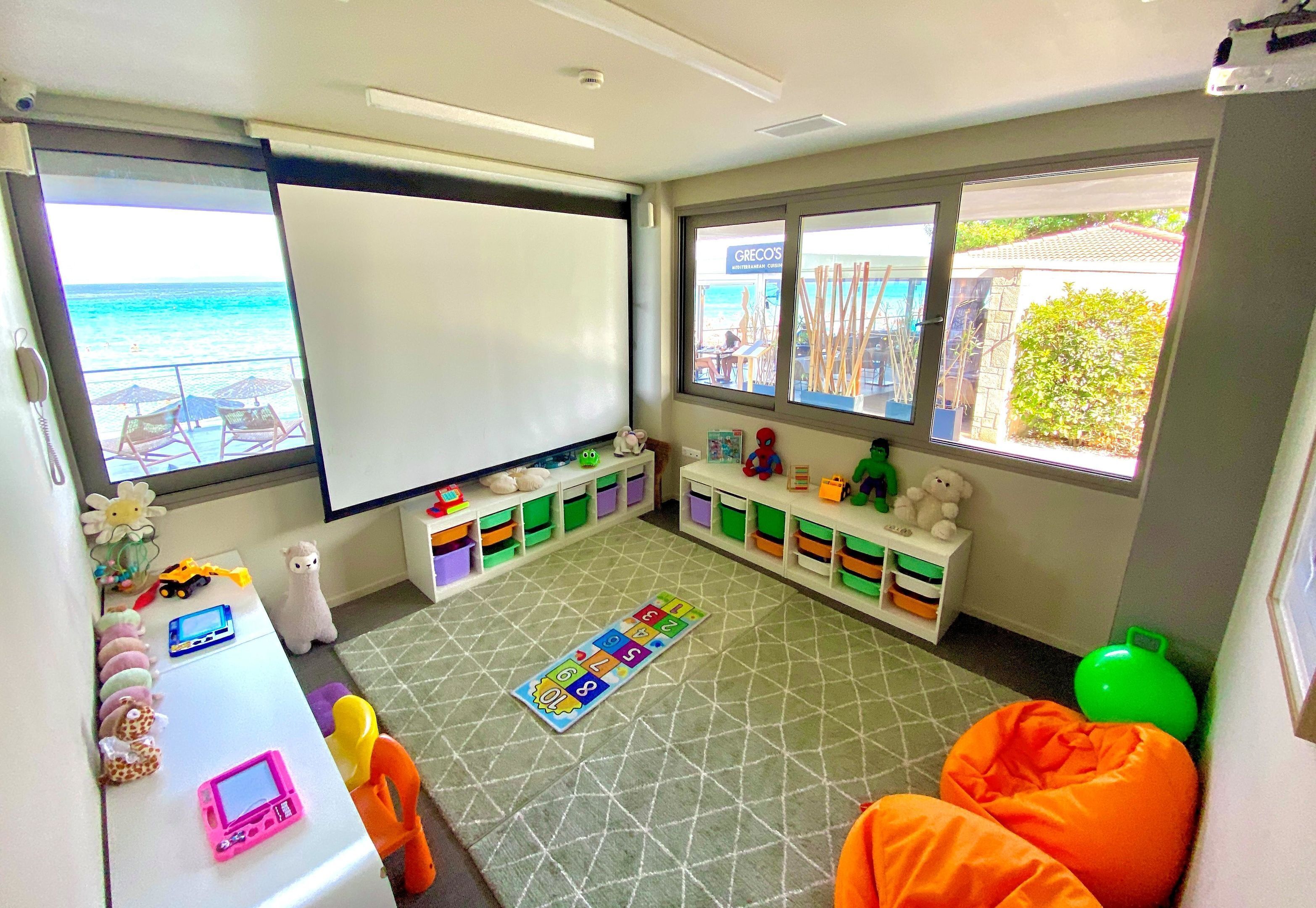 Una habitación con una pizarra, cajas de almacenamiento, un tapete de juegos y vistas a la playa.