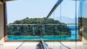 A modern interior with a large glass window offering a view of a coastal landscape with a forested hill and a body of water. - Ammon Zeus Luxury Beach Hotel (Kassandra)