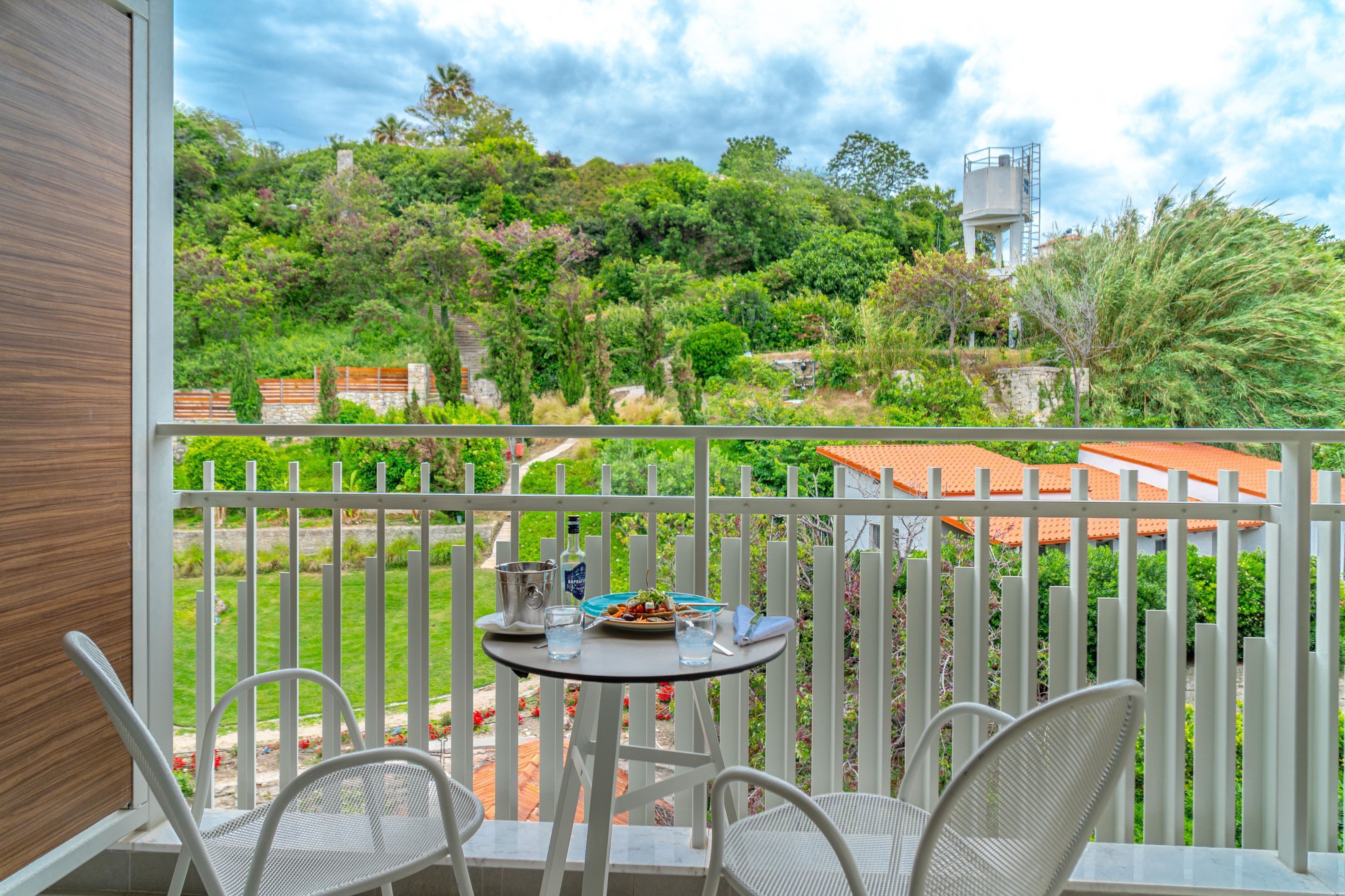 Un balcon avec une table et des chaises, donnant sur un jardin luxuriant et un château d’eau.