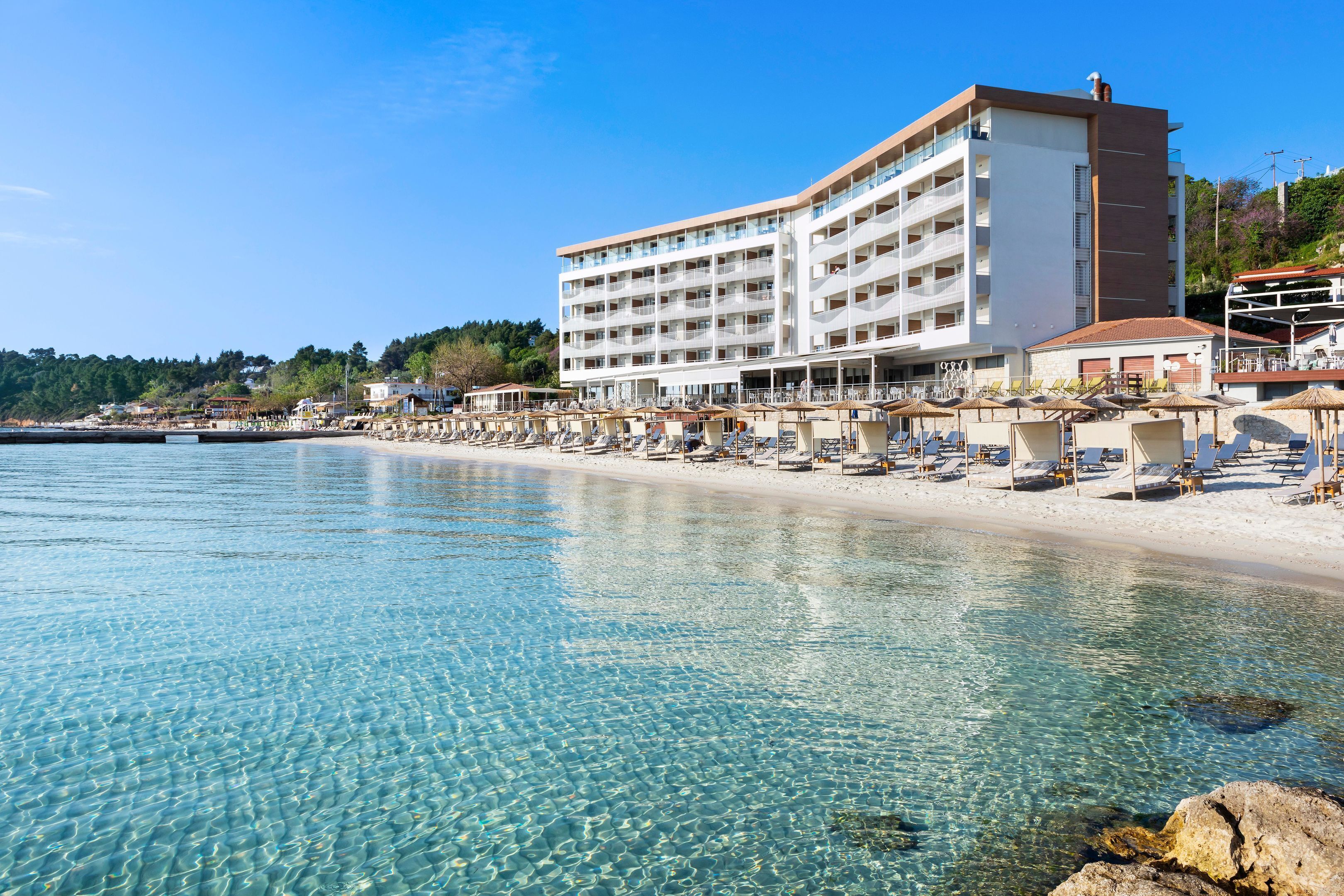 Un hôtel situé en bord de mer, avec une eau cristalline, une plage de sable et un grand bâtiment doté de nombreux balcons.