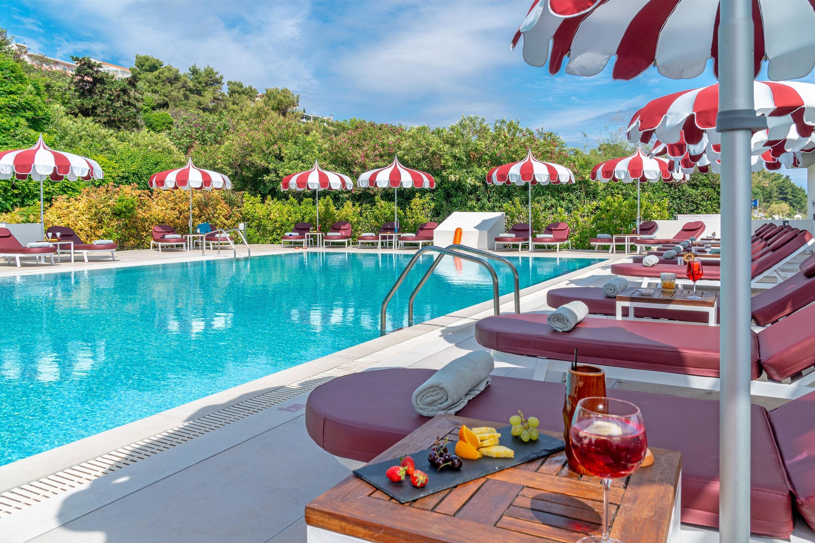 Área de piscina com guarda-sóis listrados em vermelho e branco, espreguiçadeiras e uma mesa com frutas e um copo de bebida vermelha.