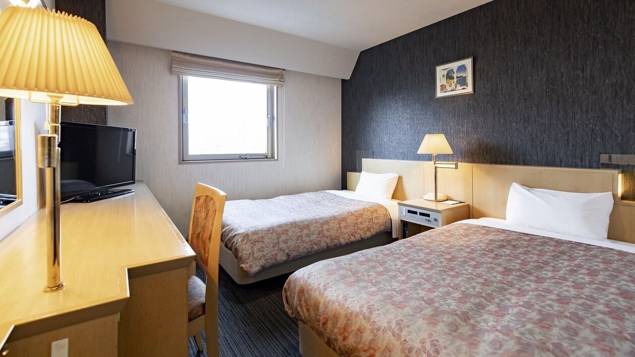 A hotel room with two beds, a desk, a TV, and a window.