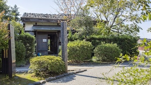 Ein traditionelles japanisches Gebäude mit einem Ziegeldach, umgeben von üppiger Vegetation und einem Kiesweg zum Eingang.