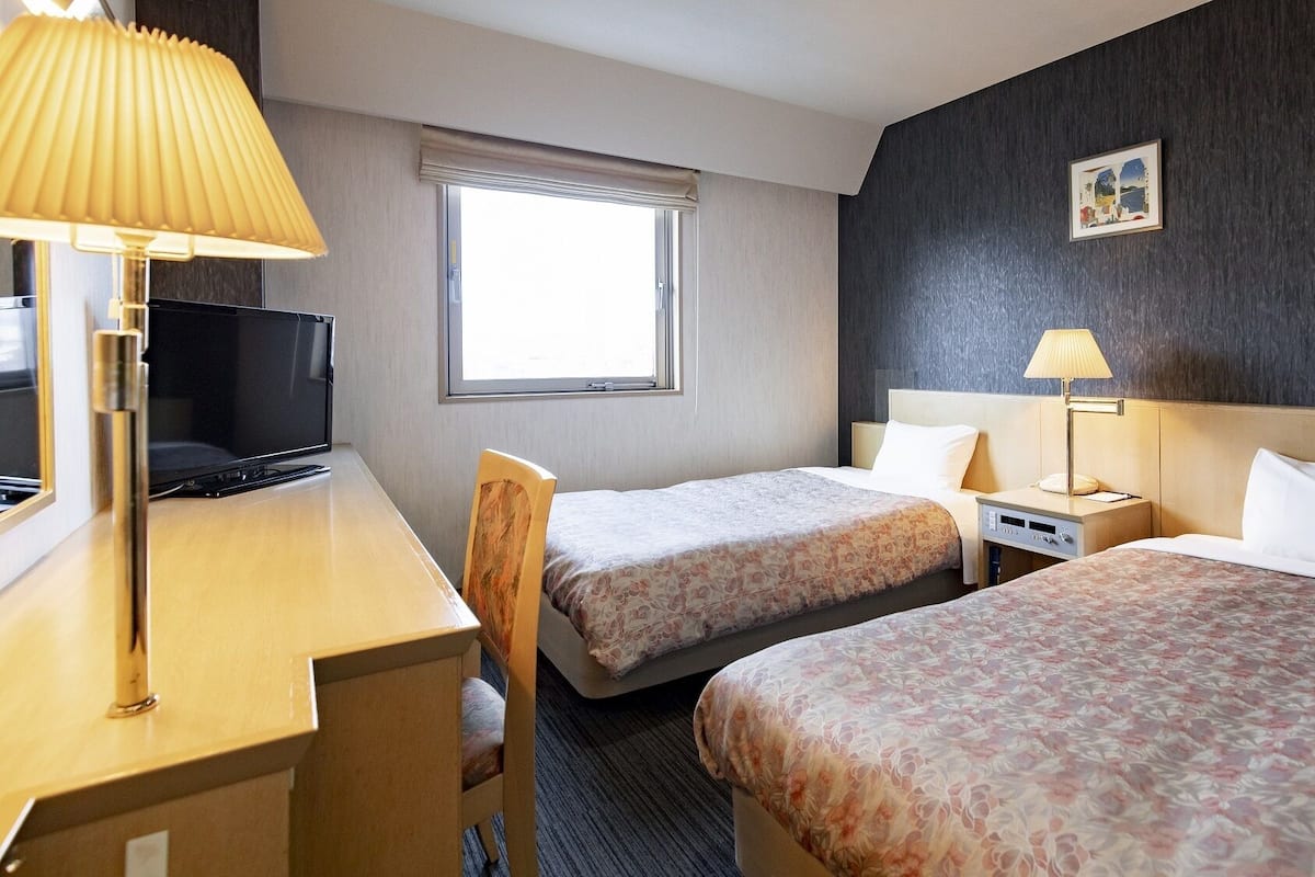 A hotel room with a desk, two beds, a TV, and a window.