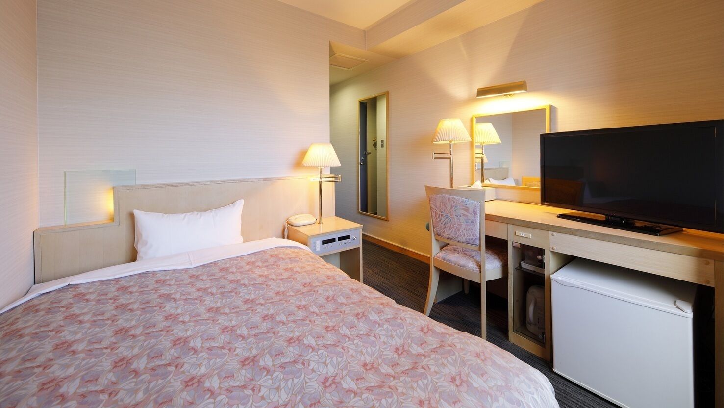 A hotel room with a bed, a desk with a television, a chair, a lamp, and a small refrigerator.