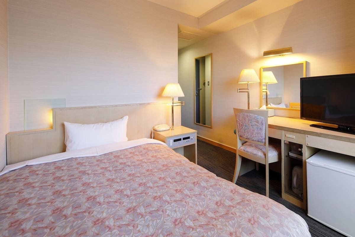 A hotel room with a bed, a desk with a television, a chair, a lamp, and a small refrigerator.