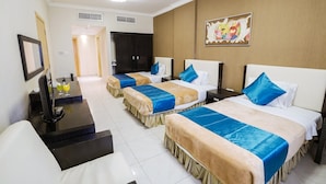 Habitación de hotel con tres camas, un escritorio con computadora y televisión. Se observa una puerta que conduce a otra habitación y un cuadro en la pared.